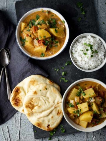 2 bowls of potato curry with a bowl of rice and flatbread (naan) on the side