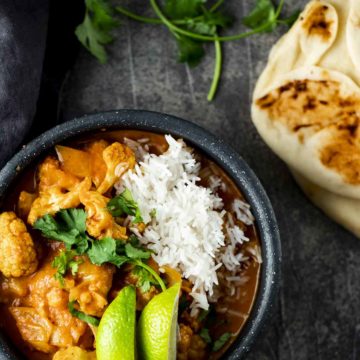 a bowl of orange cauliflower curry with white rice and lime slices and naan on the side