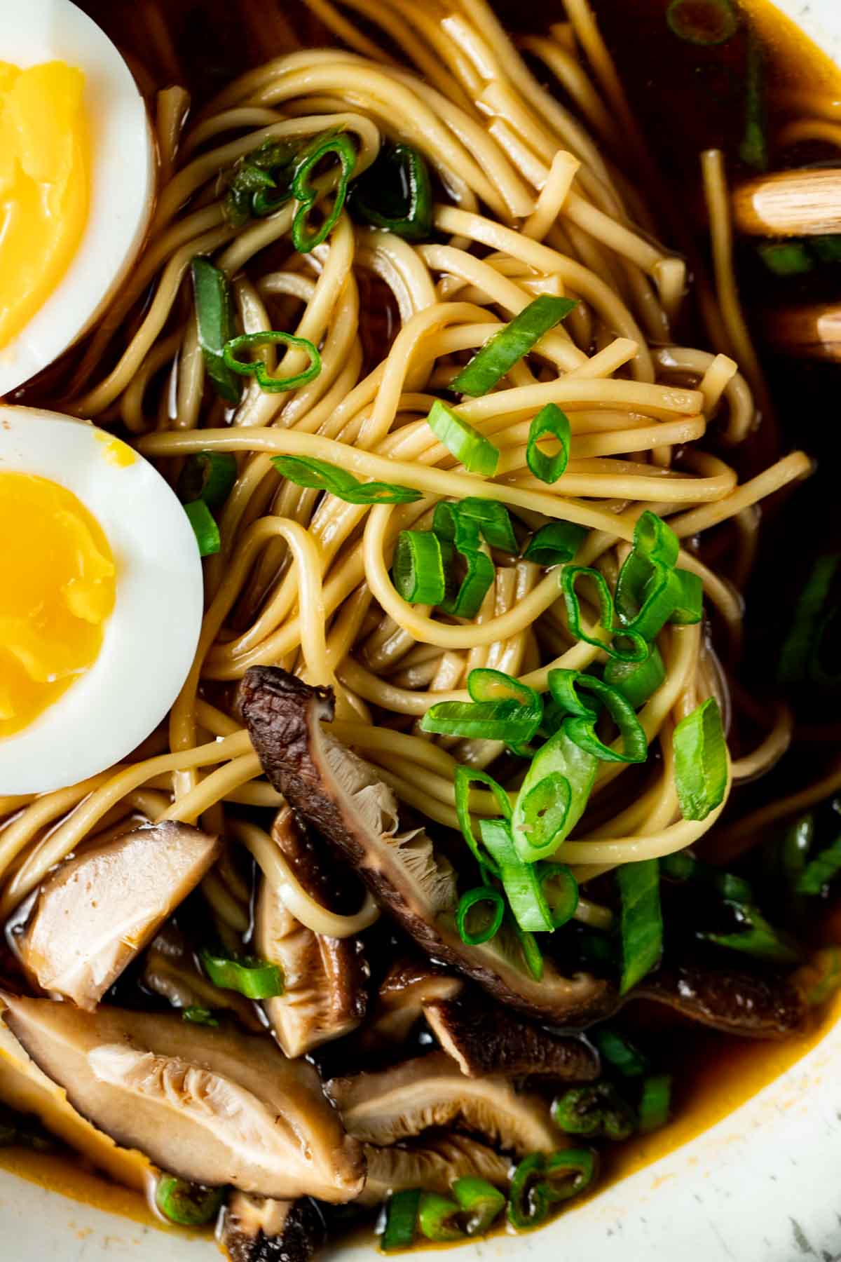 a close up bowl of dark soup with yellow noodles, green onions and sliced mushrooms in it