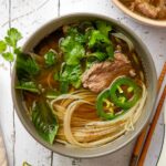 rare beef noodles soup in a bowl with green herbs and slices of pepper