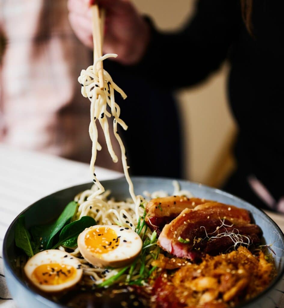 chopsticks holding noodles over a bowl of ramen with a person in the background